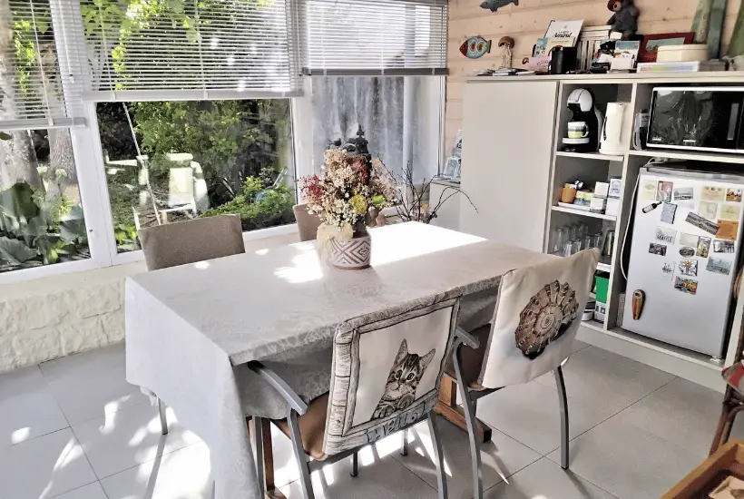 Véranda lumineuse aménagée pour le petit-déjeuner avec table, grandes fenêtres sur le jardin et réfrigérateur